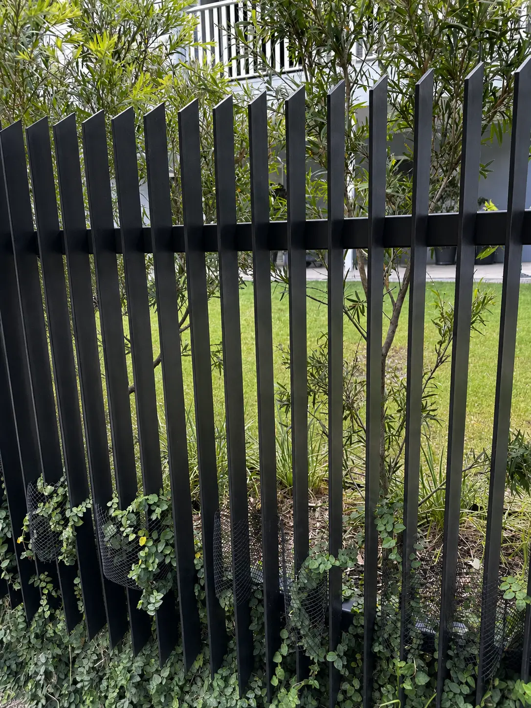 Close view of black 45 degree aluminium blade fencing beside dense residential landscaping