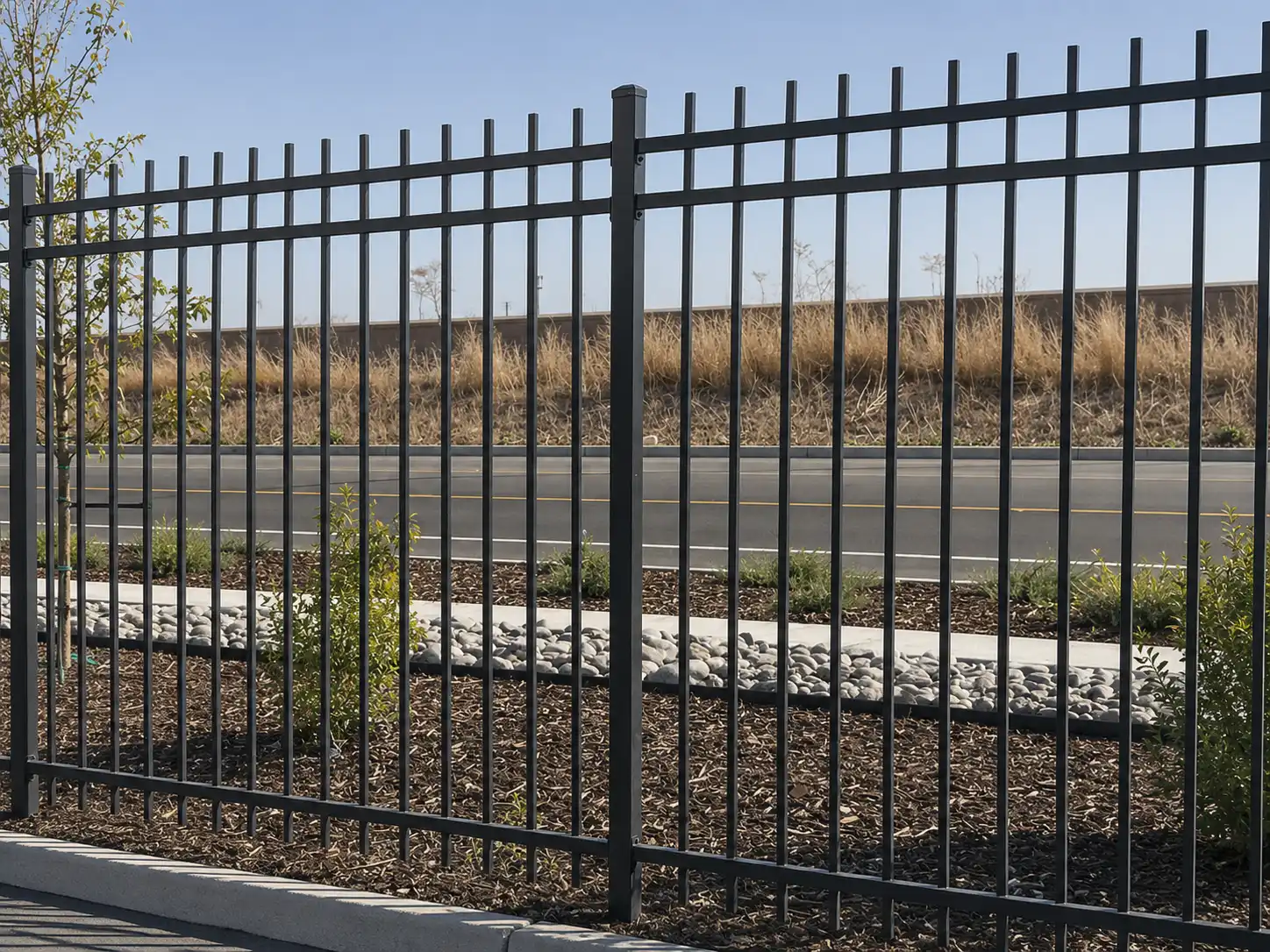 Black three-rail steel rod fence installed along a landscaped roadside boundary